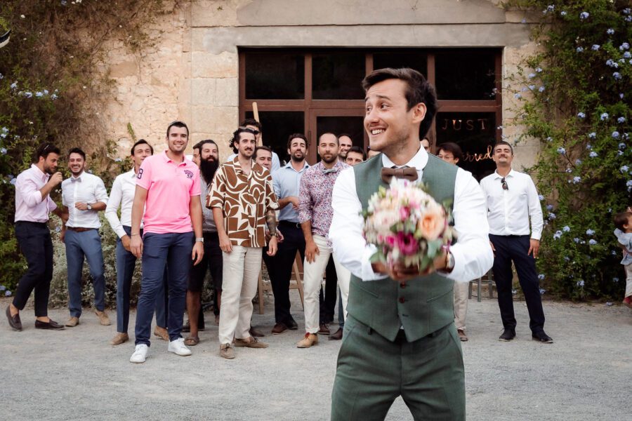 Marié prêt à lancer le bouquet à un groupe d'hommes devant une bâtisse en pierre en Occitanie